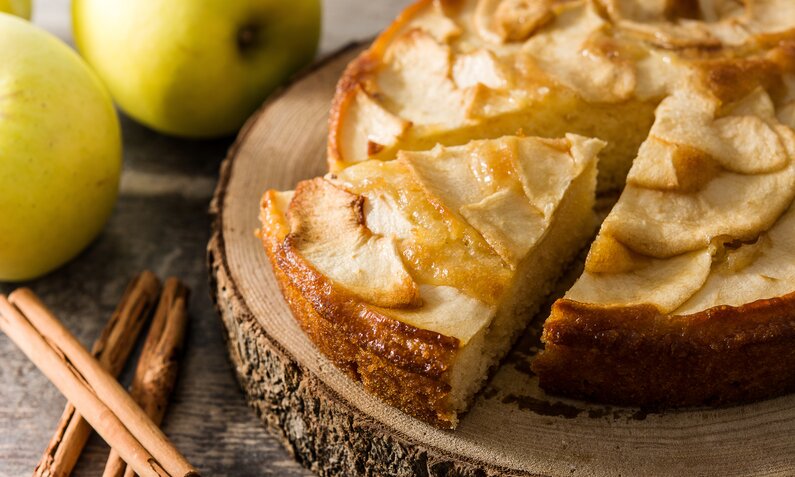 Angeschnittener Apfel-Schmandkuchen auf einem Holzbrett, daneben frische grüne Äpfel und drei Zimtstangen. | © Adobe Stock / chandlervid85