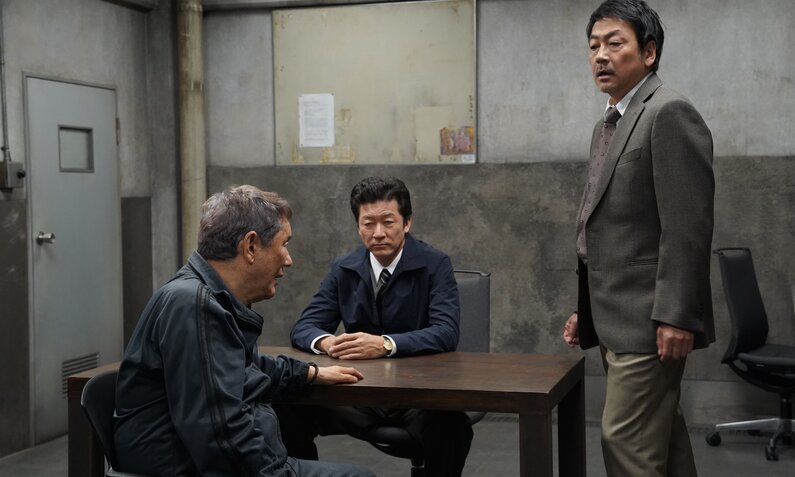 Takeshi Kitano und zwei Co-Stars sitzen und stehen an einem Tisch in einem Verhörraum in einer Szene aus dem Prime-Video-Film "Broken Rage". | © Prime Video/Amazon