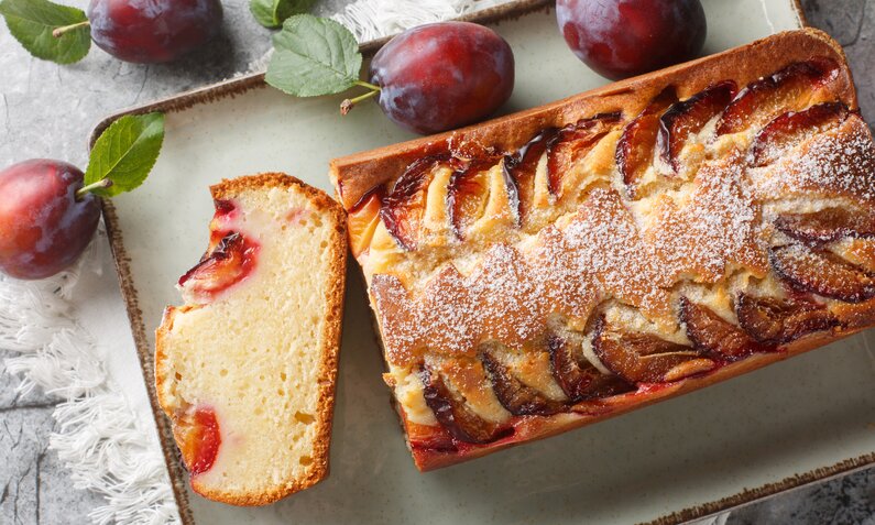 Angeschnittener Pflaumen-Joghurtkuchen von Oben auf einem Holzbrett, daneben frische Pflaumen. | © Getty Images / ALLEKO