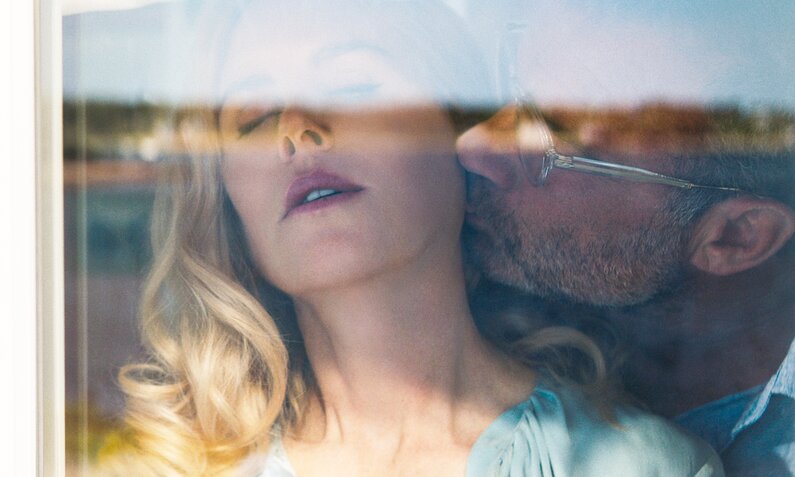 Hinter einer Fensterscheibe sieht man in einer Szene aus der Netflix-Miniserie "Ein neuer Sommer" wie Liev Schreiber Nicole Kidman auf den Hals küsst | © Netflix