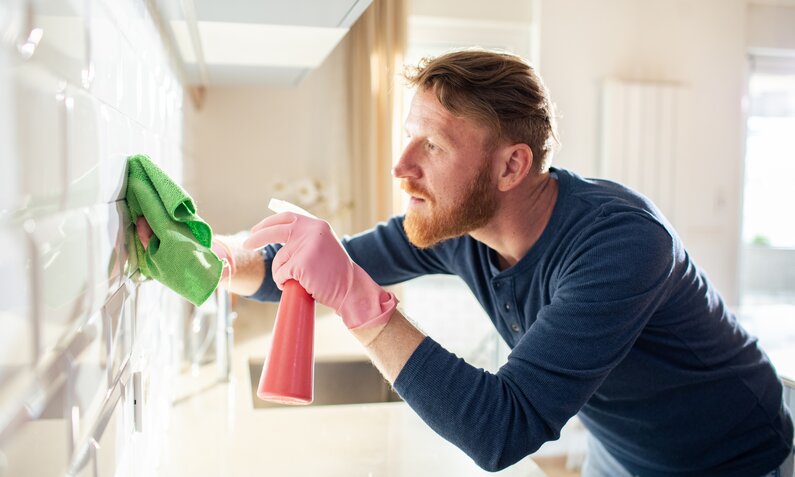 Ein Mann in blauem Pullover reinigt am Tag mit einem Spray und einem Tuch die glänzende Wand seiner Küche. | © Adobe Stock/ Vorda Berge