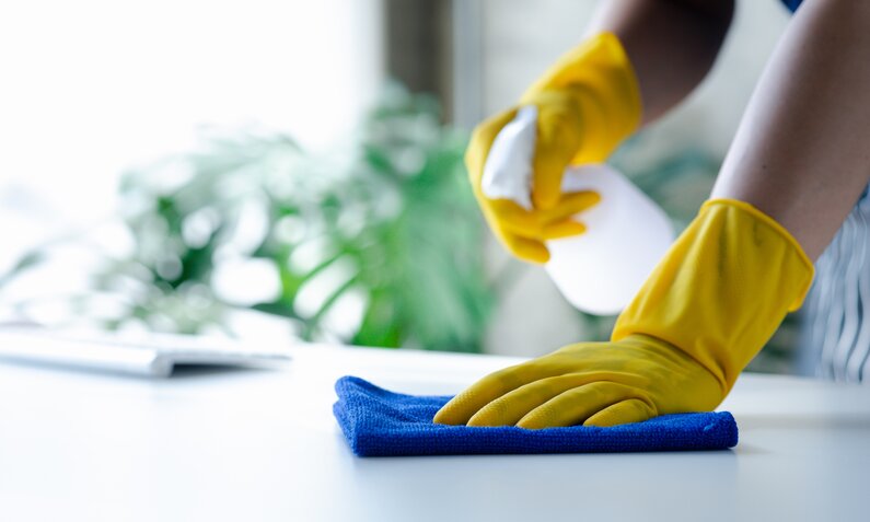 Eine Person mit gelben Handschuhen wischt mit einem blauen Tuch und einem Sprühreiniger einen Tisch ab. | © Adobe Stock/ kamiphotos