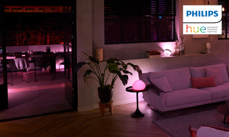 Mit Philips Hue-Lampen beleuchtetes Wohnraum mit stilvollem grauem Sofa, Pflanze und Tischlampe, Terrasse im Hintergrund in rotes Licht getaucht, gemütliche Stimmung. | © Amazon/Phlips