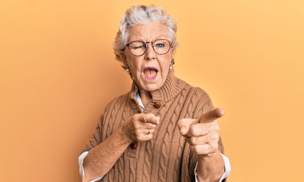 Ältere Frau mit grauen Haaren und Brille zeigt lachend mit beiden Fingern nach vorn vor orangefarbenem Hintergrund. | © Adobe Stock/ Krakenimages.com