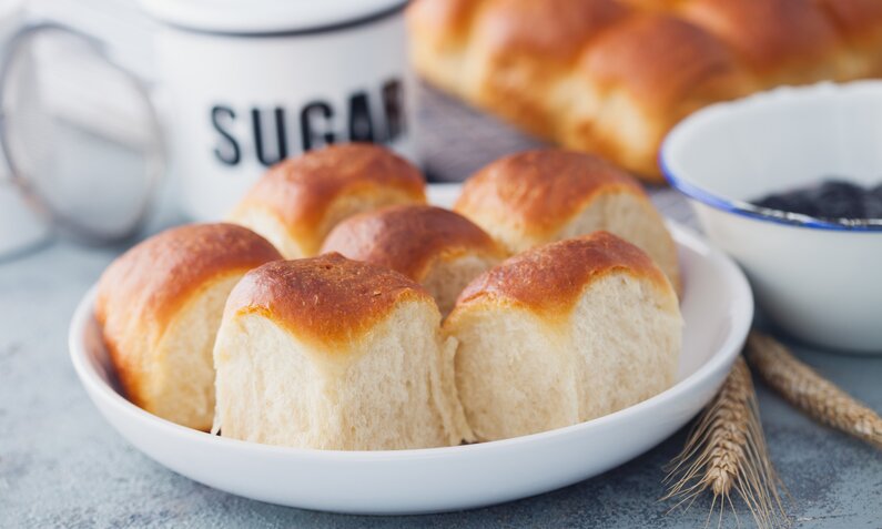 Mehrere süße Quarkbrötchen auf einem weißen Teller, dahinter Tasse mit Sugar drauf. | © Adobe Stock / matka_Wariatka