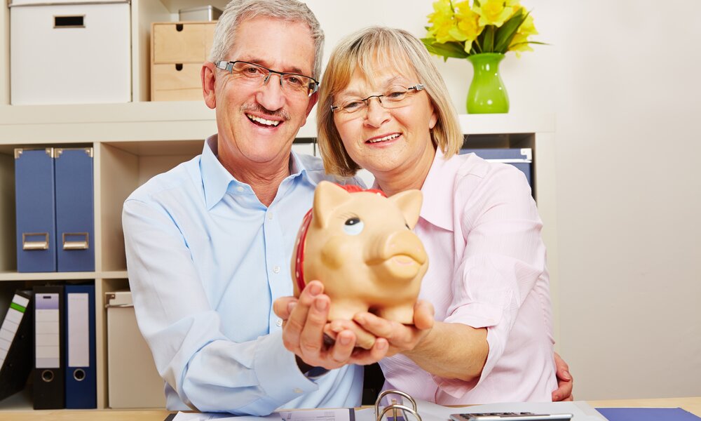 Ein älteres Paar mit Brille lächelt fröhlich in die Kamera, während es gemeinsam ein Sparschwein in einem Büro hält. | © Adobe Stock/ Robert Kneschke