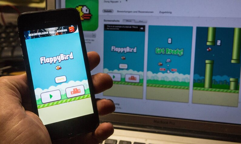 Eine Hand hält ein Smartphone, auf dem der Startbildschirm des Spiels Flappy Bird zu sehen ist, im Hintergrund läuft dasselbe Spiel auf einem Laptop. | © IMAGO / Rüdiger Wölk