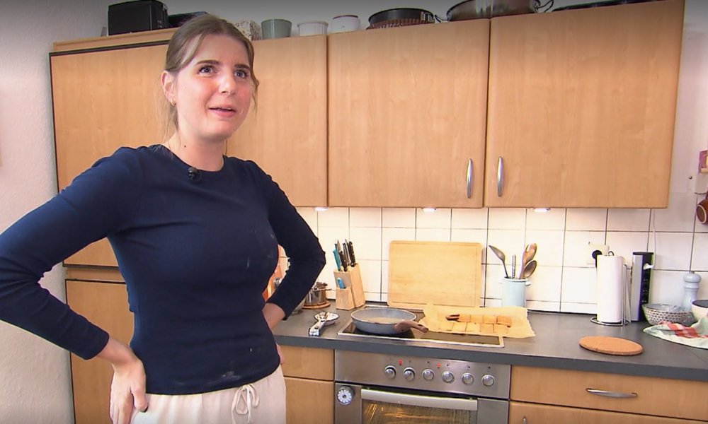 Junge Frau mit braunen Haaren im Dutt steht nachdenklich in einer Holzküche vor dem Herd, auf dem gebackene Kekse liegen. | © Screenshot Vox/RTL+ 