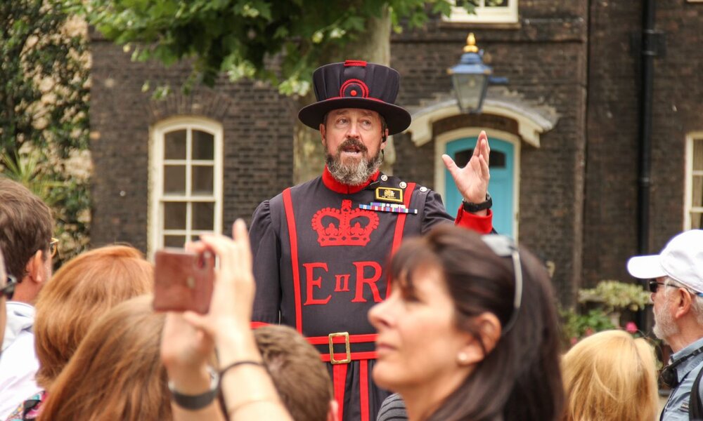 Ein Yeoman Warder in traditioneller Uniform spricht mit erhobener Hand zu einer Touristengruppe vor historischen Gebäuden. | © Getty Images / LightRocket / SOPA Images / Kontributor