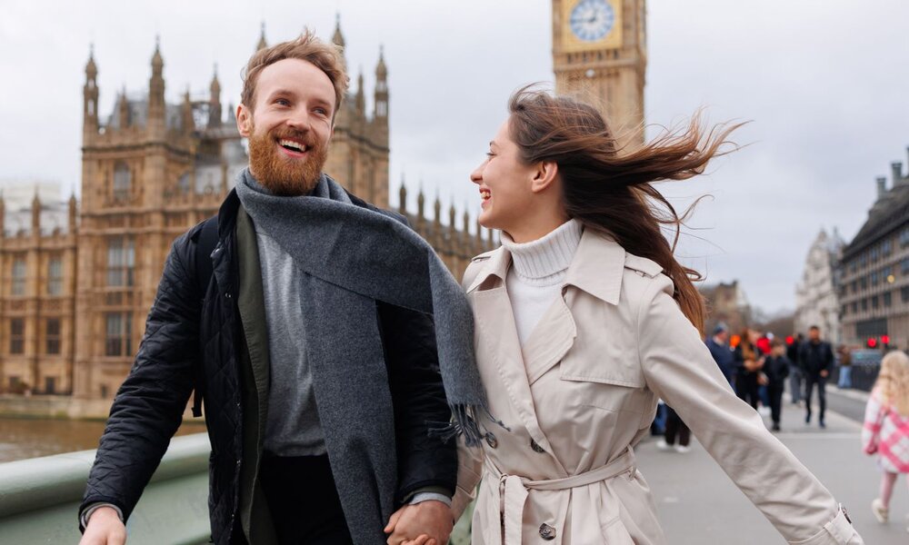 Ein lächelnder Mann mit rotem Bart und grauem Schal hält die Hand einer Frau im Trenchcoat, während sie vor Big Ben spazieren. | © Andrii  /stock.adobe.com