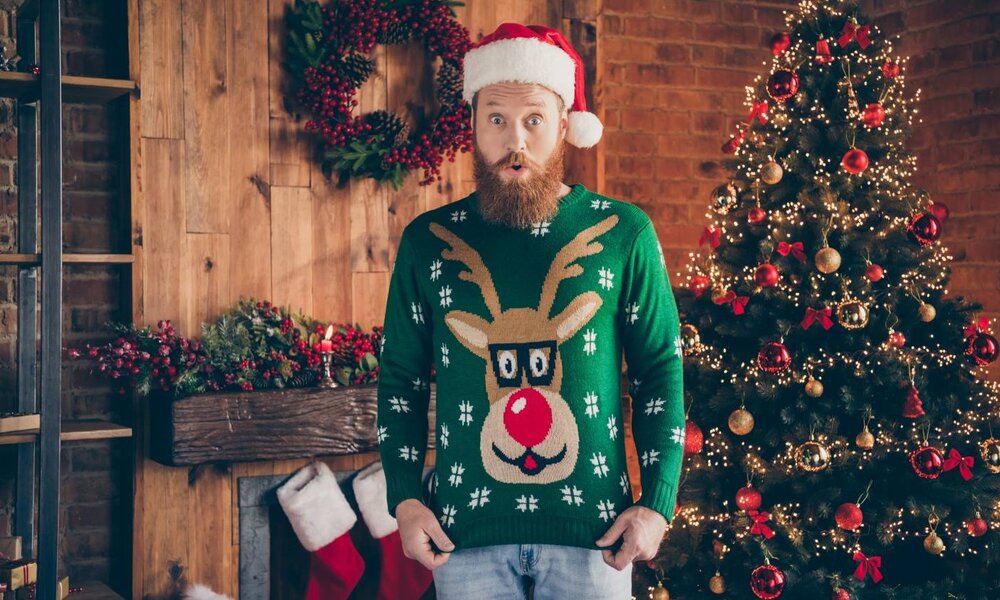 Ein bärtiger Mann in Rentierpullover und Weihnachtsmütze steht vor einem festlich dekorierten Raum mit Weihnachtsbaum und Kamin. | © deagreez/stock.adobe.com 