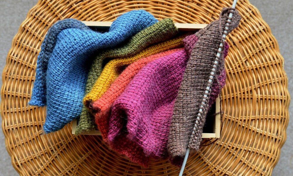 Mehrere gestrickte Stoffe in bunten Farben liegen auf einem runden Korbtisch, eine Stricknadel steckt in einem braunen Strickstück. | © Kate / stock.adobe.com