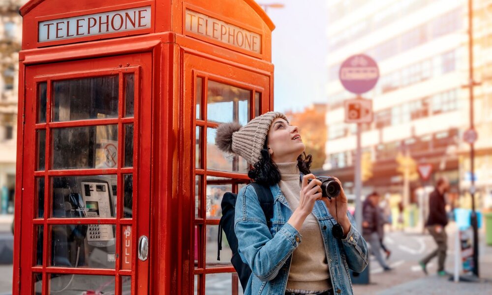 Touristin mit Rucksack und Kamera steht lächelnd vor einer roten Telefonzelle, während Menschen im Hintergrund vorbeigehen. | © Iryna/stock.adobe.com