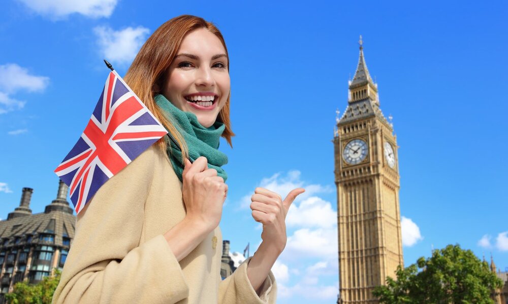 Junge Frau mit langen braunen Haaren hält eine britische Flagge vor dem Big Ben und lächelt glücklich in die Kamera. | © ryanking999/stock.adobe.com