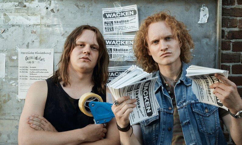 Sebastian Jakob Doppelbauer als Thomas Jensen und  Sammy Scheuritzel als Holger Hübner in der RTL+ Serie "Legend of Wacken". Die beiden Gründer des Heavy Metal Festivals sind beide isind im Metal-Look der Neunziger mit Denim und schwarzen T-Shirts, blicken entschlissen und halten Flyer fürs festival hoch. | © RTL+/Thomas Leidig