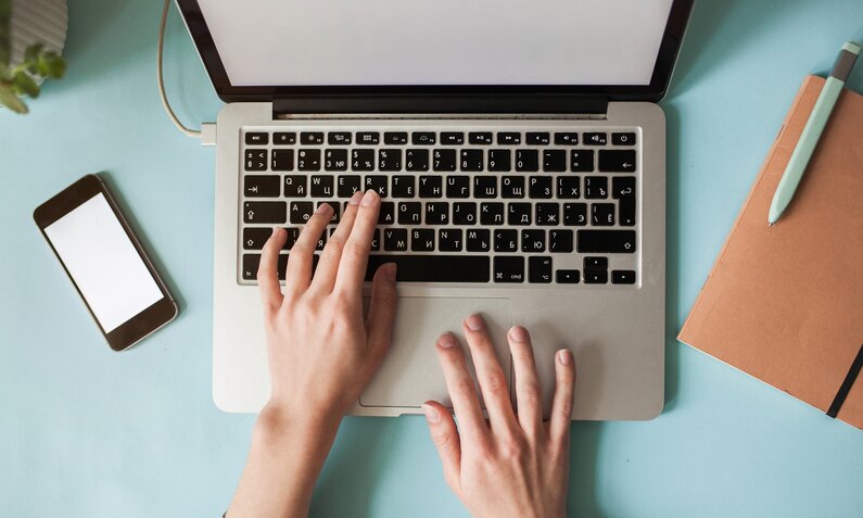 Frauenhände auf einer Laptop-Tastatur aus der Vogelperspektive. Links auf dem Tisch neben dem Computer liegt ein Smartphone. | © Getty Images/Igor Ustynskyy