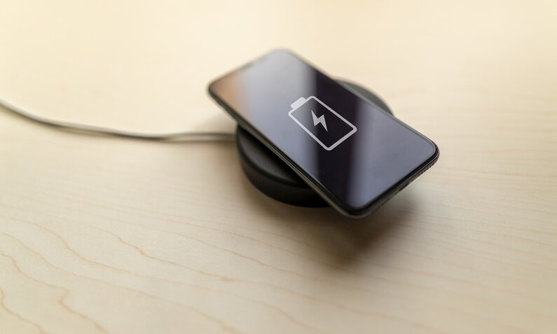 Ein schwarzes Smartphone liegt in einer schwarzen Ladeschale. Die steht auf einem hellen Holztisch. | © Getty Images/Javier Zayas Photography