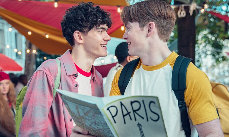 Joe Locke und Kit Connor als Nick und Charlie in einer Szene der Netflix-Serie "Heartstopper", Staffel 2. Die beiden stehen auf der Straße und strahlen sich an, Kit hält einen Paris-Reiseführer | © Netflix/Teddy Cavendish