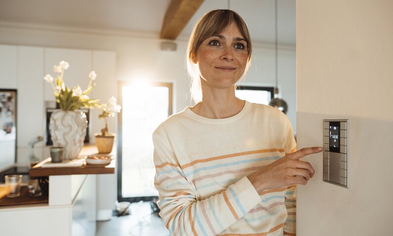 Frau lächelt, während sie an einem Thermostat in der Wand Einstellungen vornimmt. | © Getty Images/Westend61