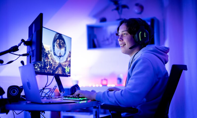 Ein junger Mann sitzt vor seinem COmputer und hat Spaß beim Spielen. Die ganze Szenerie ist in blaues Licht getaucht. | © Getty Images/ Alistair Berg