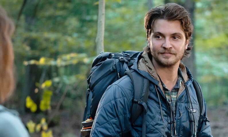 Ellie Kemper als Helen Luke Grimes als Jake in der Netflix-Filmromanze "Küssen und andere lebenswichtige Dinge". Jake dreht sich zu Helen um, er trägt blaue Wanderkleidung und steht vor einem grünen Waldhintergrund. | © Netflix/Barbara Nitke