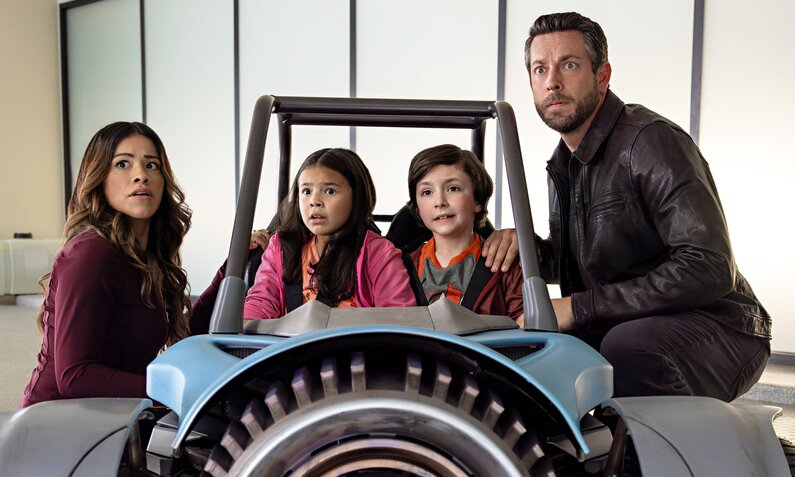 Gina Rodriguez als Nora Torrez, CEverly Carganilla als Patty Torrez, Connor Esterson als Tony Torrez und Zachary Levi als Terrence Torrez im Netflix-Film "Spy Kids: Armageddon". Die beiden Kinder sitzen in einem Spielauto in einem leeren Raum, ihre Eltern sitzen rechts und links von ihnen. | © Netflix/Robert Rodriguez