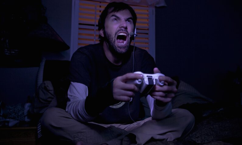 Ein Mann mit einem Controller in der Hand sitzt in einem dunklen Raum und schreit vor Wut. | © Getty Images/Romona Robbins Photography