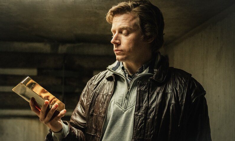 Jack Lowden in einer Szene der BBC-Serie "Das Gold". Er trägt eine Lederjacke, steht in einem Keller oder einer Garage und hält einen Goldbarren in der Hand, auf dessen polierter Oberfläche sich sein Gesicht spiegelt. | © Paramount+