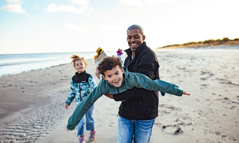 Ein Mann tobt mit seinem Sohn am Strand herum, beide lächeln fröhlich, ein Mädchen läuft links hinter dem Mann hinterher. | © Getty Images/Marko Geber