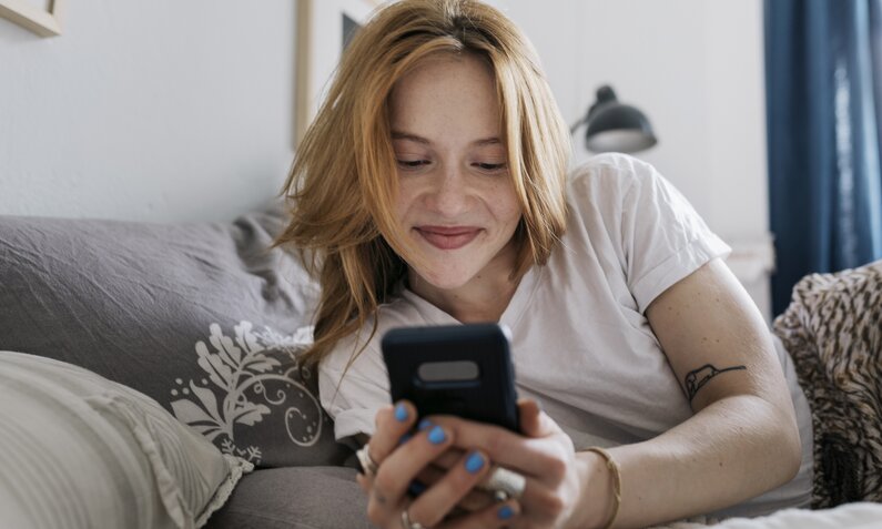 Junge Frau lächelt, während sie, auf dem Sofa liegend, auf ihr Smartphone sieht. | © Getty Images/Willie B. Thomas
