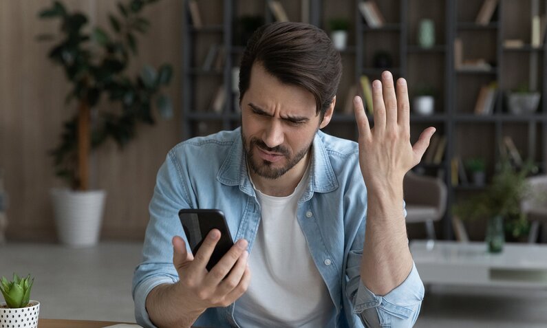 Mann sitzt am Tisch und ärgert sich über etwas, das er auf seinem Smartphone sieht. | © Getty Images/fizkes