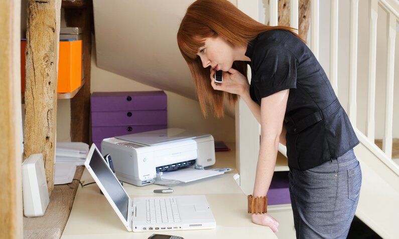 Eine junge rothaarige Frau steht im Profil am Schreibtisch, auf dem neben einem Computer auch ein Drucker steht. | © Getty Images/Tim Robberts