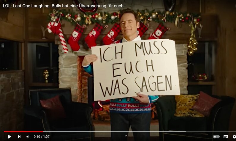 Michael Bully Herbig steht mit Weihnachtspulli in einem festlich dekorierten Wohnzimmer und hält Schrifttafeln hoch. Auf der obersten steht "Ich muss Euch was sagen" | © Screenshot YouTube/Amazon Prime Video