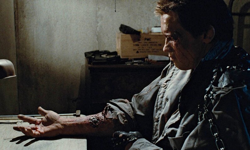 Arnold Schwarzenegger sitzt in einer Szene des Kinofilms "The Terminator" in einem dunklen Raum an einem Tisch und starrt auf seinen Arm in dem die technik des Roboterkillers sichtbar wird, den er spielt | © ddp images/ LMKMEDIA Ltd.
