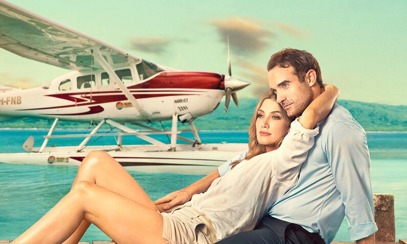 Delta Goodrem und Joshua Sasse sind auf der Key Art zum Netflix Film "Love is in the Air" aneinander gelehnt auf einem Anlegesteg sitzend zu sehen. Im Hintergrund ist türkisblaues Meer, blauer Himmel und ein Wasserflugzeug zu sehen. | © netflix