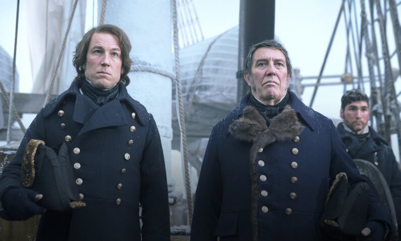 Ciaran Hinds und eine Co-Star stehen in einer Szene aus der Serie "The Terror" in Offiziersuniformen an Deck und blicken ernst | © 2017 AMC Film Holdings LLC. All Rights Reserved.