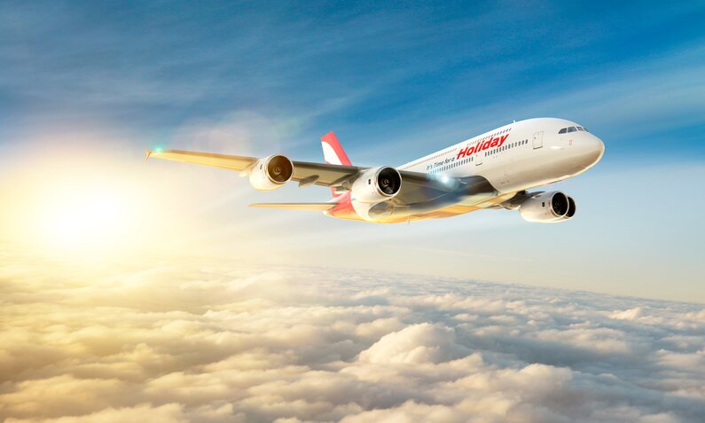 Ein Flugzeug im Flug knapp über den Wolken im Licht der Sonne - auf der Seite steht in roter Schrift "Holiday". | © Getty Images/Aaron Foster