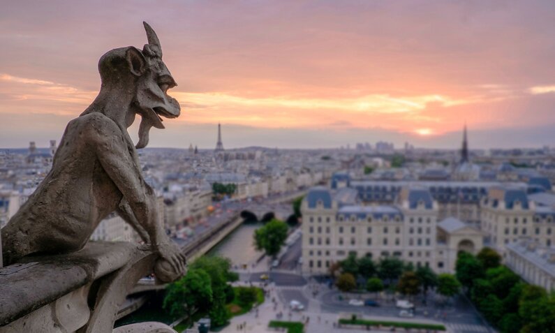 Im Vordergrund ein steinerner ziegenähnlicher Gargoyle auf einem Dach, im Hintergrund ein Panoramablick über die Stadt Paris mit Eiffeltum bei Sonnenuntergang | © Unstplash/Pedro Lastra