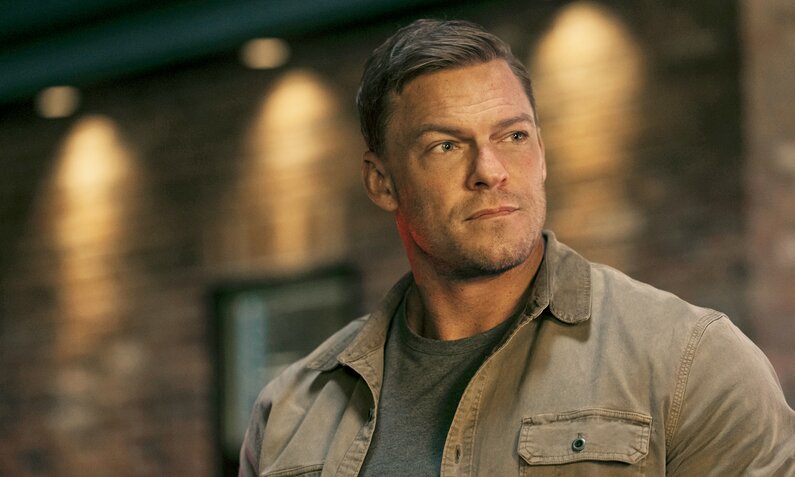 Nahaufnahme von Alan Ritchson als Jack Reacher in der Prime Video Serie "Reacher". Er steht bei Nacht draußen, im Hintergrund ist eine beleuchtete Backsteinmauer zu sehen.  | © Prime Video