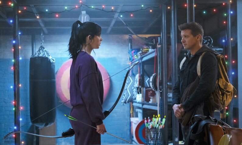 Hailee Steinfeld und Jeremy Renner stehen sich in einem abgedunkelten Raum mit großen regalen und bunten Minilichterketten gegenüber in einer Szene aus der Marvel Serie "Hawkeye" | © Marvel Studios 2021. All Rights Reserved.