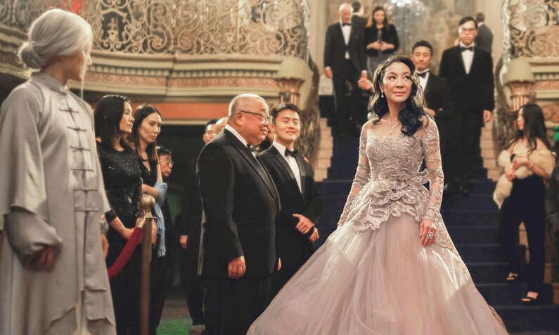 Michelle Yeoh steht im Abendkleid inmitten festlich gekleideter Menschen in einem prächtigen Raum mit großer Treppe in einer Szene aus dem Kinofilm "Everything Everywhere All At Once". | ©  ddp images /LMKMEDIA Ltd.