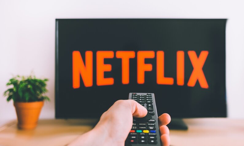 Ein großer Fernseher mit großem Netflixlogo steht auf einem Holzboard mit Blumentopf an der Seite vor einer weißen Wand, im Vordergrund ist eine Hand mit TV-Fernbedienung zu sehen. | © Unsplash/freestocks