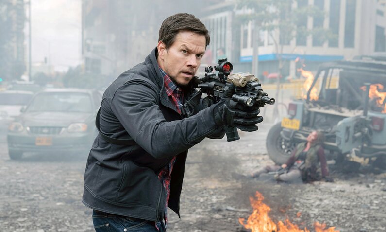 Mark Wahlberg steht in einer Szene des Kinofilms "Mile 22" mit Gewehr im Anschlag auf einer Straßenkreuzung, im Hintergrund sind Feuer, beschädigte Autos zu sehen. | © ddp imges/ Murray Close