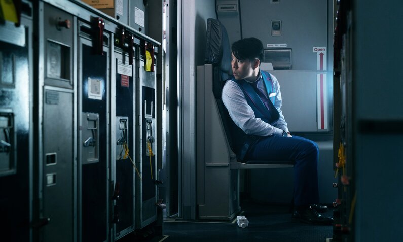 Jeremy Ang Jones sitzt als Flugbegleiter in einer Szene der Apple TV+ Serie "Hijack" im Flugzeug auf einem Sitz am Ende der Flugzeugküche. es ist düster und er ist allein. | © Apple TV+