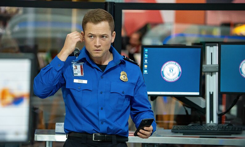 Taron Egerton in blauer Sicherheitsbeamtenuniform vor Computermonitoren in einer Szene aus dem Netflix-Film "Carry-on". Er hält ein Smartphone in der einen Hand, mit der anderen setzt er einen Kopfhörer auf. | © Netflix/Sam Lothridge