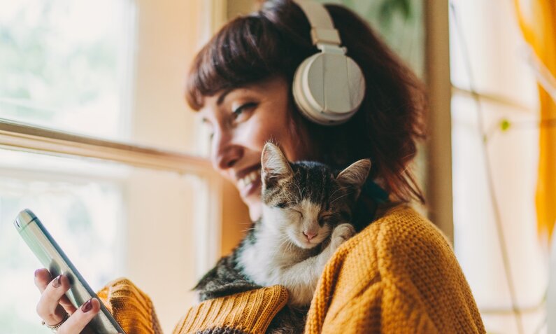 Frau mit Katze hört Podcast | © Getty Images / E+ / martin-dm
