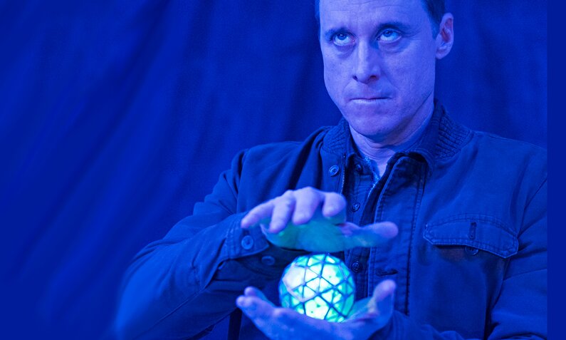 Alan Tudyk steht in blauem Licht und hält eine leuchtendweiße Kugel in der Hand in einer Szene der Serie "Resident Alien" | ©  ddp Images/Capital Pictures