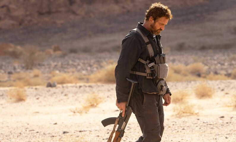 Gerard Butler geht mit gesenktem Kopf und Gewehr in der Hand durch eine karge Landschaft in einer Szene aus dem Kinofilm "Kandahar" | © Imago Images/Landmark Media