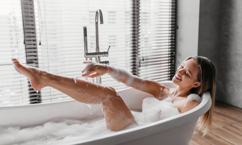 Frau, die in einer Badewanne sitzt und sich die Beine rasiert  | © Adobe Stock/ Prostock-studio