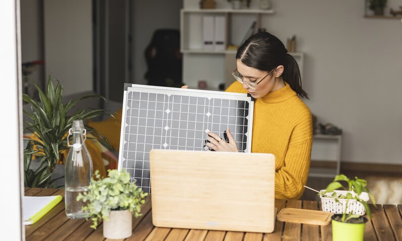 Eine Frau sitzt am Schreibtisch und hält ein kleines Solarmodul in der Hand. | © Getty Images/Westend61